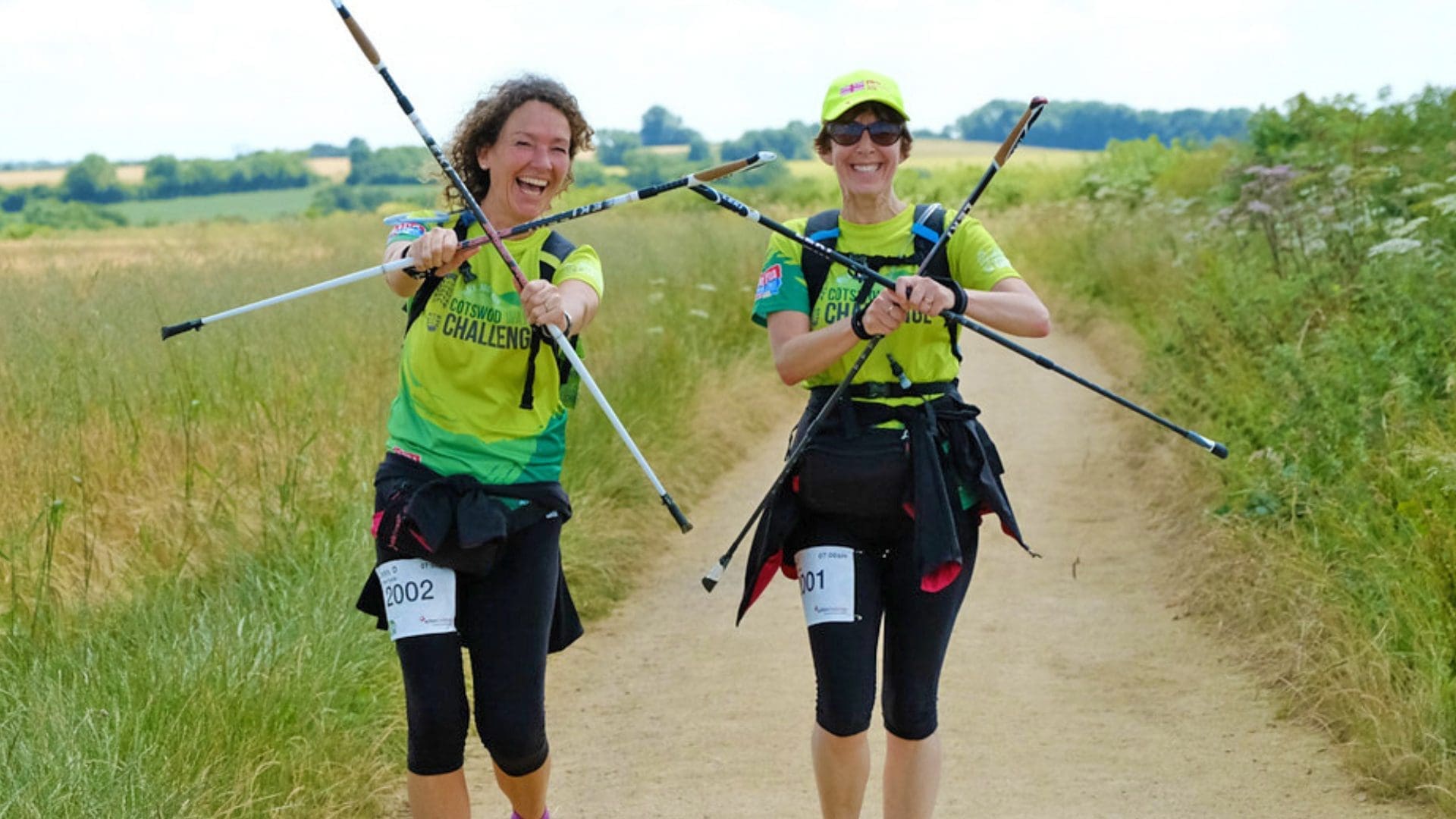 Two women smiling as the walk down a pathway in the countryside. They are making crosses using their hiking sticks