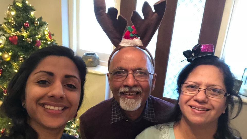 Grown up daughter standing next to her father and her mother. They are standing in front of a Christmas tree. The father is wearing reindeer antlers