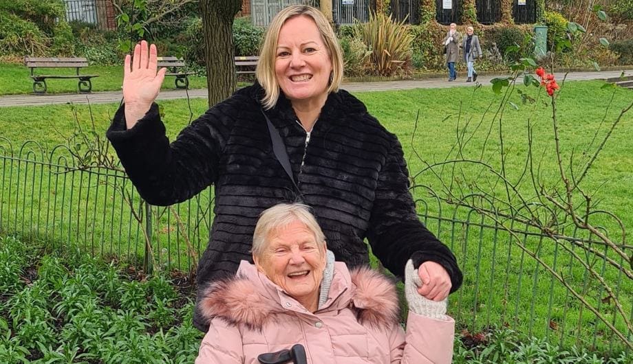 Daughter standing smiling and waving behind her elderly mother who is wheelchair bound smiling and holding her daughter's hand whilst sitting in the foreground