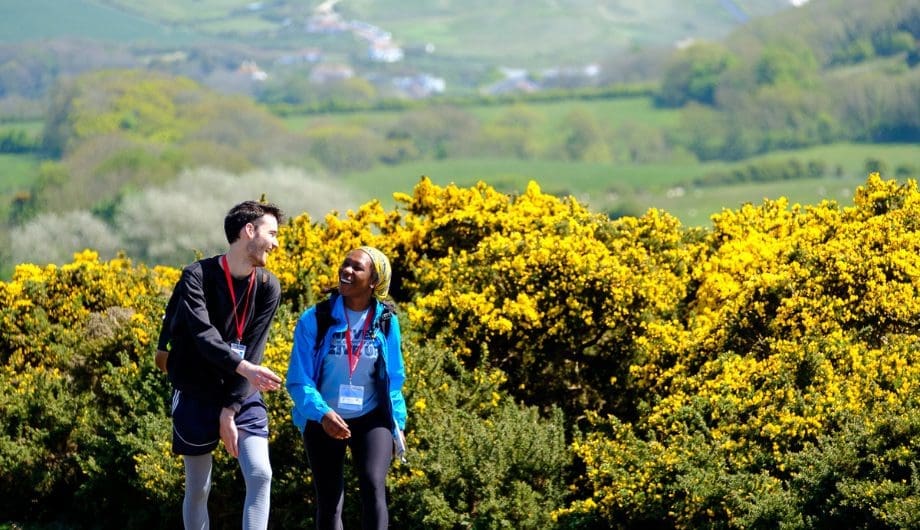 Man and woman talking and laughing together while walking. Behind them is a countryside view and hedges of flowers