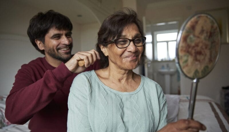 A man (carer) helps a woman to get ready in the bedroom in the morning.
