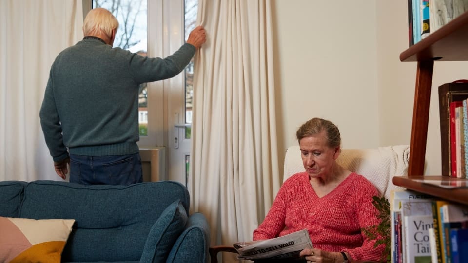 elderly male and femal couple. The elderly man draws the curtains closed while the elderly woman is sitting reading the newspaper