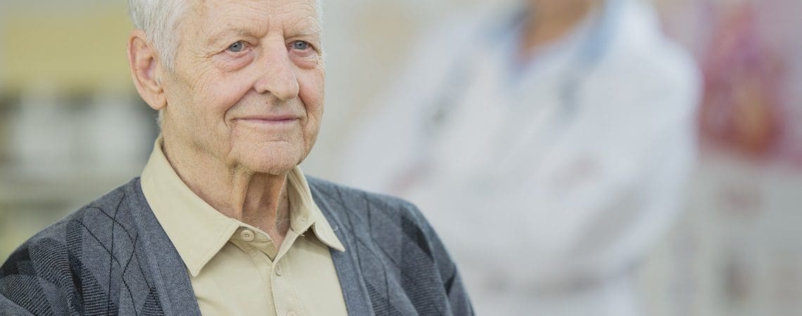 A Caucasian female doctor and elderly Caucasian man are indoors in a hospital. She is wearing doctor's clothes and he is wearing casual clothing. The man is in focus with the woman behind him.