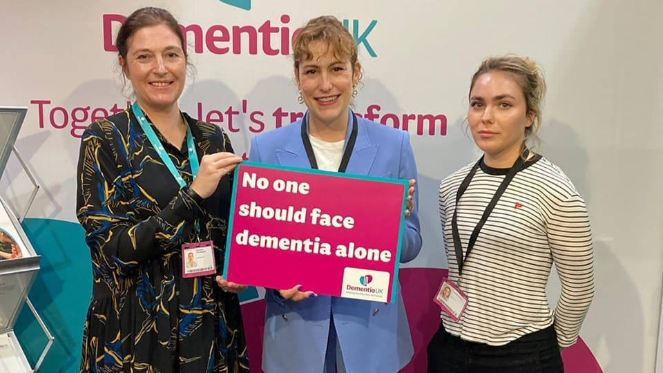 MP Victoria Atkins holding sign that reads no one should face dementia alone. Standing next to Faradane and Connie from our Campaigns team