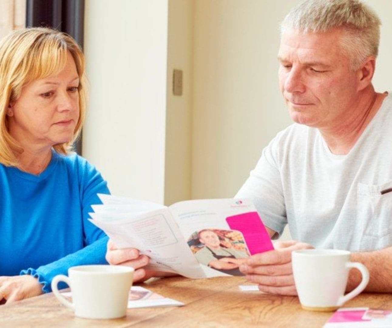 Husband and wife sat at the dining table reading a Dementia UK leaflet with two mugs of tea