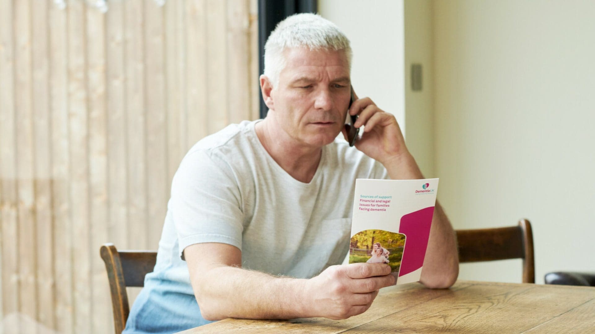 man sat at the dining table on the phone whilst looking at a Dementia UK leaflet